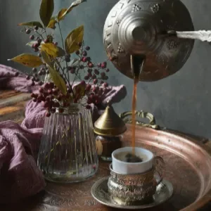 Teko logam antik sedang menuangkan kopi ke dalam cangkir keramik bermotif di ata nampan tembaga dengan dekorasi bunga.